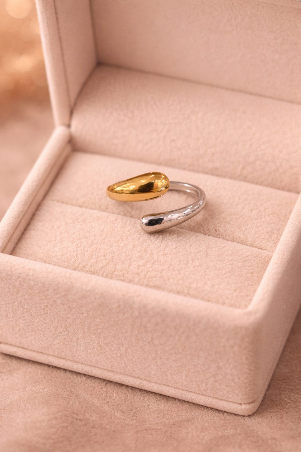 Divu toņu atvērta tipa gredzens ar vienu zeltainu un vienu sudrabotu noapaļotu galu, novietots uz gaišas samta rotu kastītes siltā, maigi izpludinātā apgaismojumā