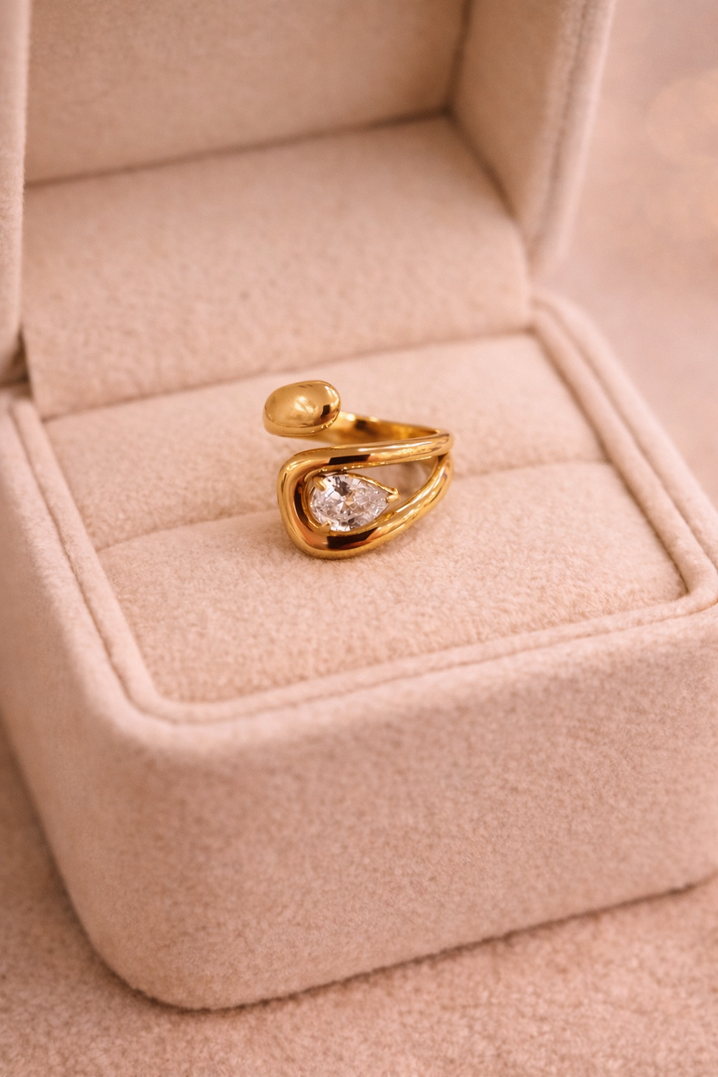 Zelta krāsas atvērta dizaina gredzens ar plūstošu, skulpturālu formu un caurspīdīgu lāsītes formas kristālu, novietots uz gaišas samta rotaslietu kastītes ar siltu, maigu apgaismojumu.