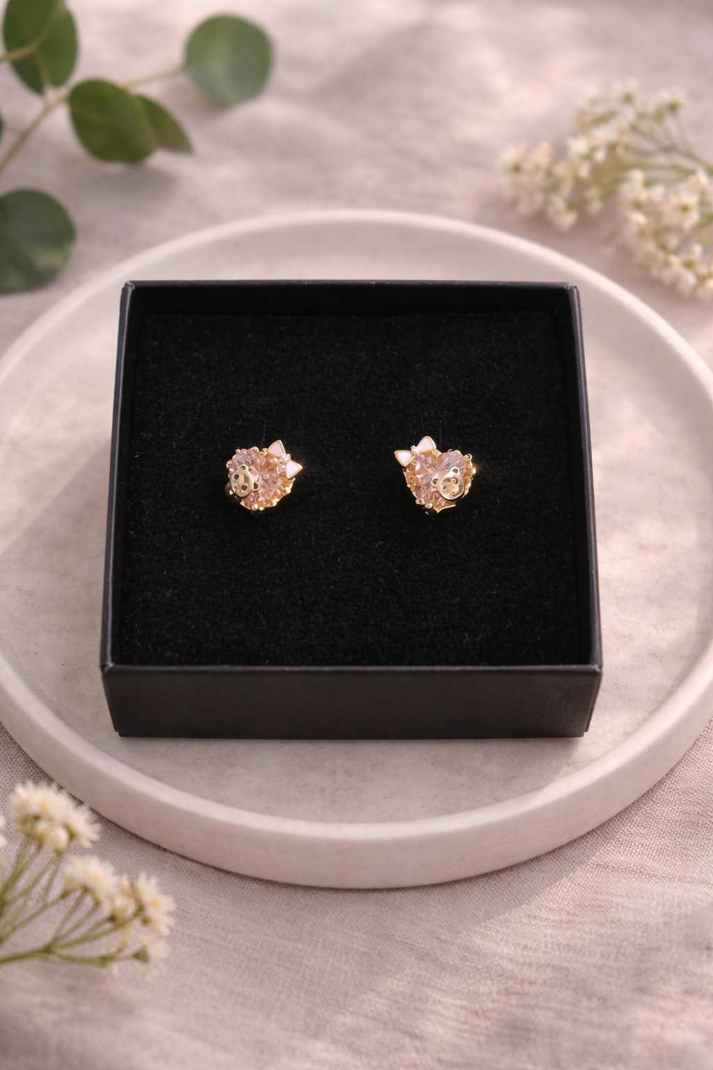 Zelta krāsas bērnu auskari ar rozā kristāliem, ievietoti melnā rotaslietu kastītē uz gaiša keramikas šķīvīša, minimālistiska rotaslietu produktu fotogrāfija ar dabīga lina fonu un maigu dienasgaismu.