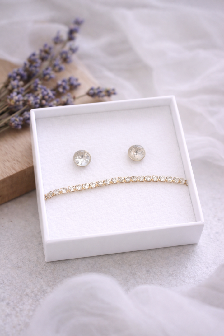 Balta rotaslietu kastīte ar baltu samta iekšpusi, kurā izvietoti apaļi, gaiši mirdzoši auskari un smalka zelta tenisa stila rokassprādze ar dzidriem akmentiņiem; fonā gaiša matēta virsma, koka dēlītis ar žāvētu lavandu un viegli izpludināts caurspīdīgs audums.