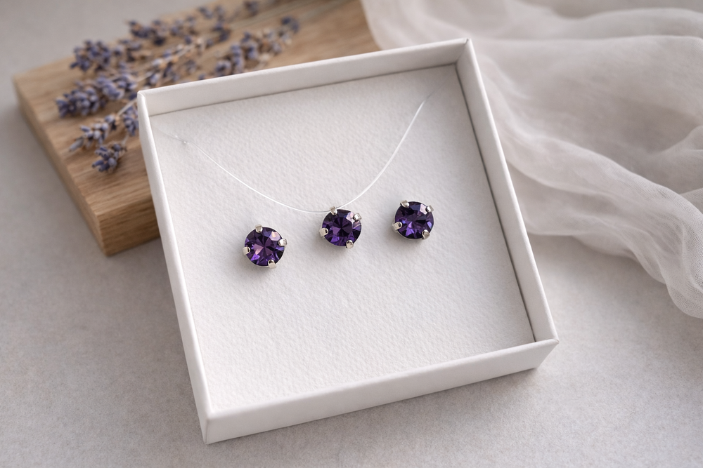 Minimālistiska rotu komplekta produktu fotogrāfija baltā kastītē – sudraba ietvarā iestrādāti violeti kristāli . Komplektā nagliņu auskari un kaklarota uz caurspīdīgas auklas. Fons gaišs, ar koka dēlīti, žāvētu lavandu un viegli izpludinātu audumu, maigs dabīgais apgaismojums.