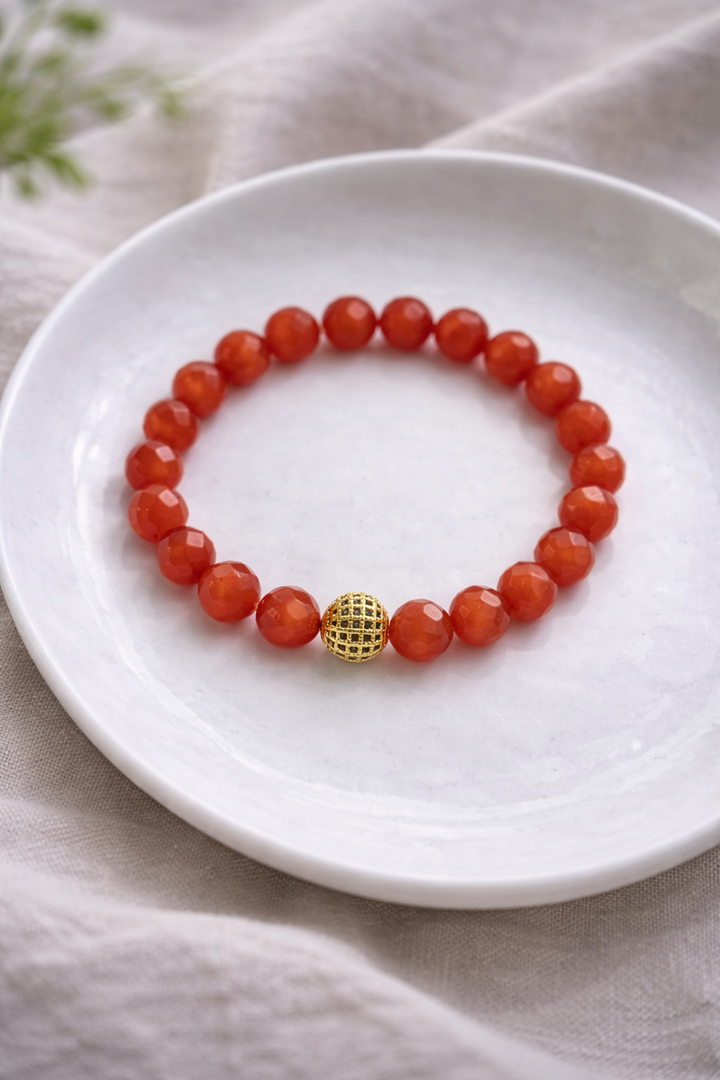 Dabīgo akmeņu aproce ar oranžām, slīpētām kaķacs pērlēm un vienu zelta krāsas dekoratīvu akcenta pērlīti centrā. Aproce simetriski izkārtota uz balta keramikas šķīvīša, fons – gaišs lina audums ar maigi izpludinātu zaļu augu elementu. Maiga, dabīga dienasgaisma izceļ akmeņu spīdumu un faktūru.