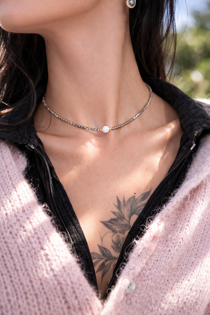 Tuvplāna fotogrāfija saulainā dienā ar sievietes kaklu un atslēgas kaulu līniju fokusā. Ap kaklu – smalks čokeris no pelēkām kristāla pērlītēm ar centrālo baltu mēnessakmens bumbiņu. Silta saules gaisma izceļ ādas mirdzumu un akmens spīdumu, redzams ziedu tetovējums uz krūtīm, fons maigi izpludināts.