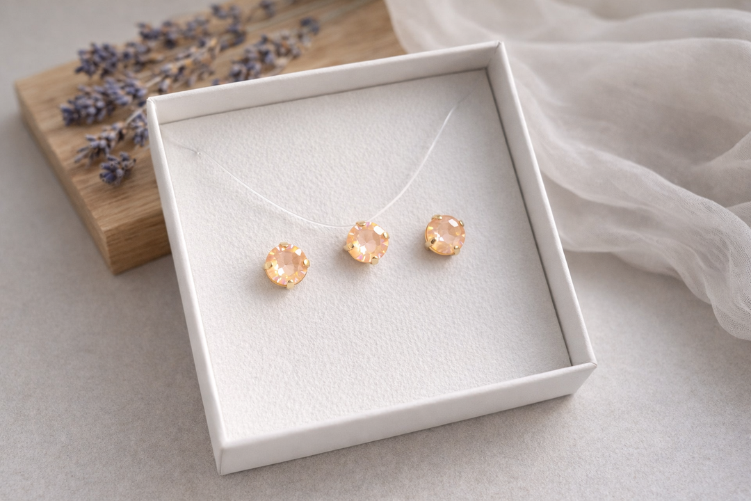 Minimālistiska rotu komplekta produktu fotogrāfija baltā kastītē – zeltītā ietvarā iestrādāti persiku krāsas kristāli.Komplektā nagliņu auskari un kaklarota uz caurspīdīgas auklas. Fons gaišs ar koka dēlīti, žāvētu lavandu un viegli plīvojošu caurspīdīgu audumu, maigs dabīgais apgaismojums un sekls asuma dziļums.
