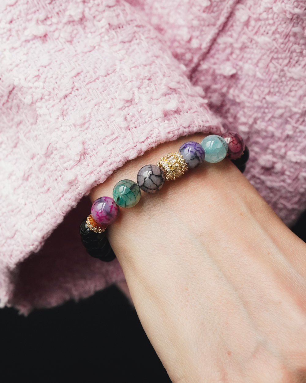 Rokassprādze ar daudzkrāsainu pūķa ahātu, melnu oniksu un zelta krāsas detaļām uz rokas pie rozā tvīda žaketes piedurknes.