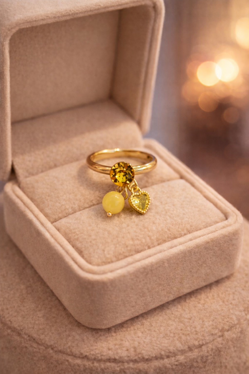 Zelta gredzens ar apaļu dzeltenu kristālu un diviem piekariņiem (gaiši dzeltenu lodīti un mazu sirds formas kulonu), novietots atvērtā gaišā samta juvelierizstrādājumu kastītē, silts apgaismojums un izpludināts fons.