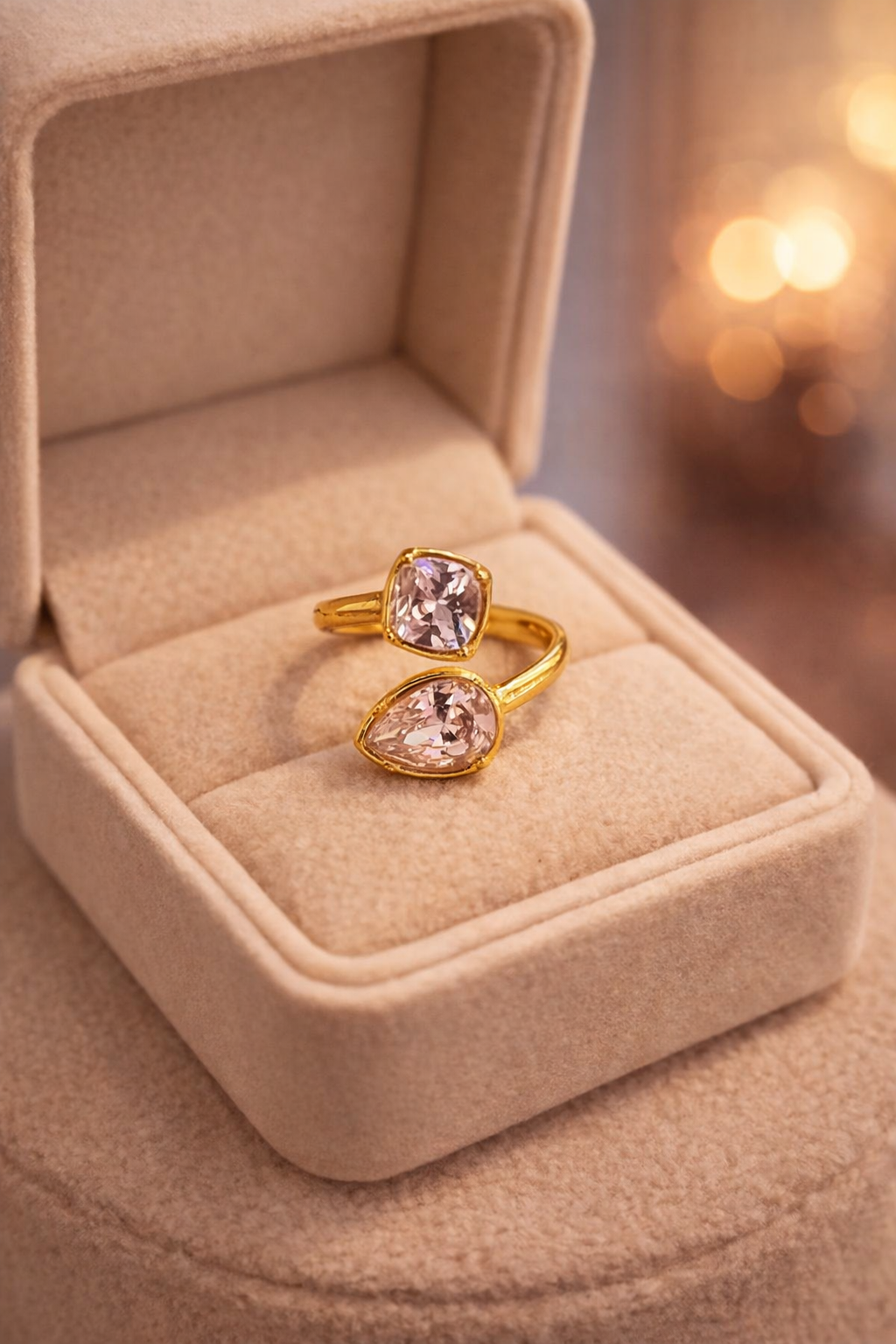 Zelta krāsas atvērta tipa gredzens ar diviem kristālbaltiem kristāliem – vienu kvadrātveida un otru lāsītes formā, novietots uz gaišas samta rotaslietu kastītes ar siltu, izpludinātu fonu.