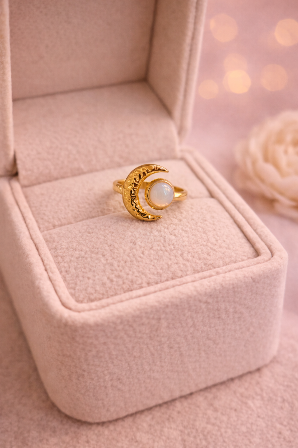 Zelta krāsas gredzens ar dekoratīvu pusmēness motīvu un apaļu, pienaini mirdzošu akmeni, novietots uz gaišas samta juvelierizstrādājumu kastītes ar siltu, izplūdušu gaismas fonu.