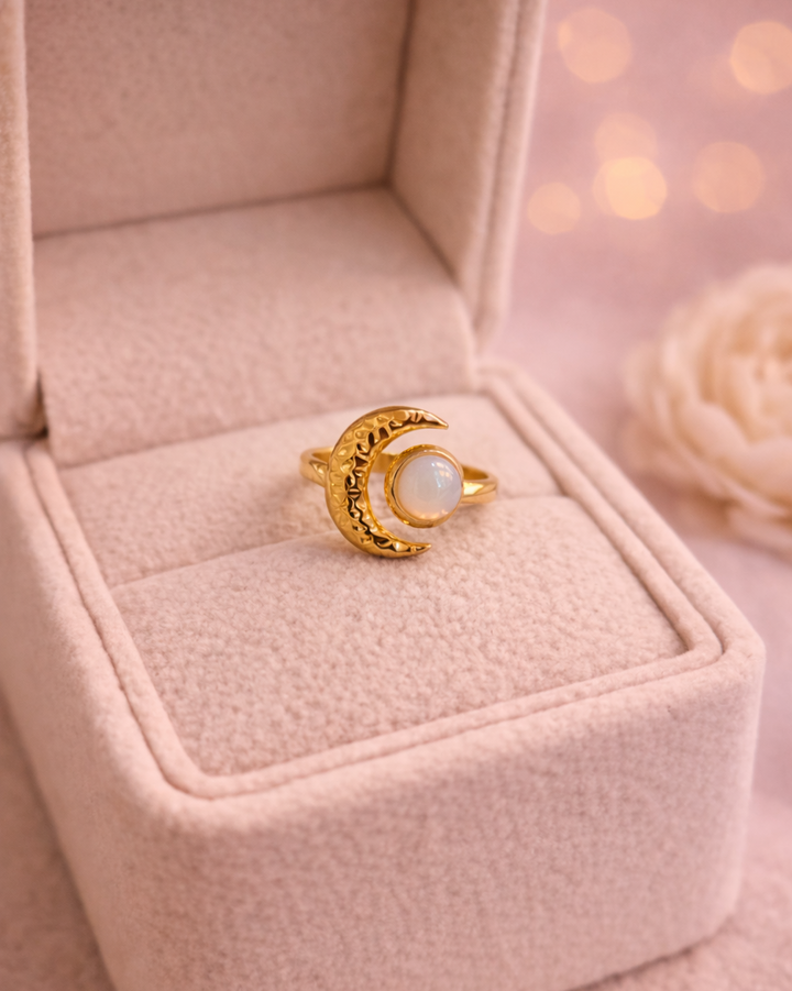 Zelta krāsas gredzens ar dekoratīvu pusmēness motīvu un apaļu, pienaini mirdzošu akmeni, novietots uz gaišas samta juvelierizstrādājumu kastītes ar siltu, izplūdušu gaismas fonu.