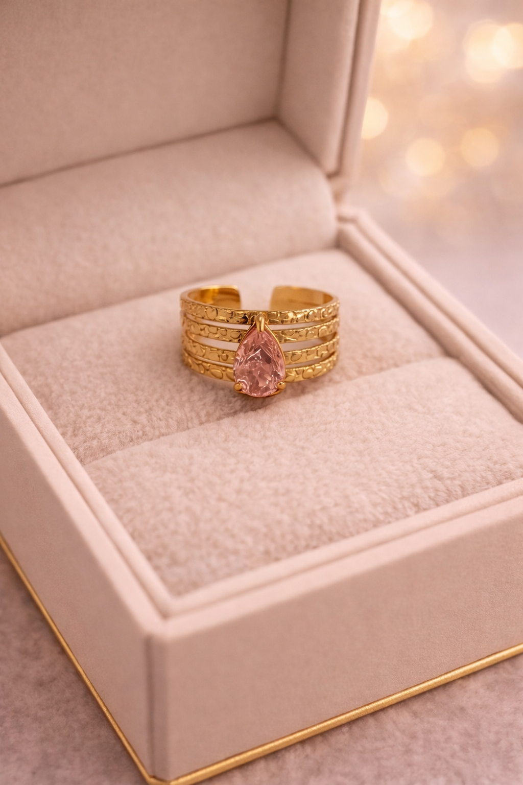 Zelta toņa daudzslāņu gredzens ar lāsītes formas rozā kristālu, novietots atvērtā gaiša samta rotaslietu kastītē ar maigu, siltu apgaismojumu un izpludinātu fonu.