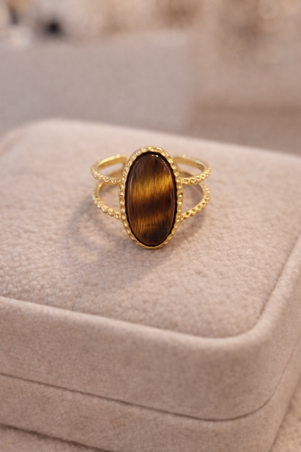 Zelta krāsas gredzens ar dubultu stīpiņu un ovālu brūnu tīģeracs akmeni, novietots uz gaišas samta juvelierizstrādājumu kastītes, maigs silts apgaismojums un izpludināts fons.