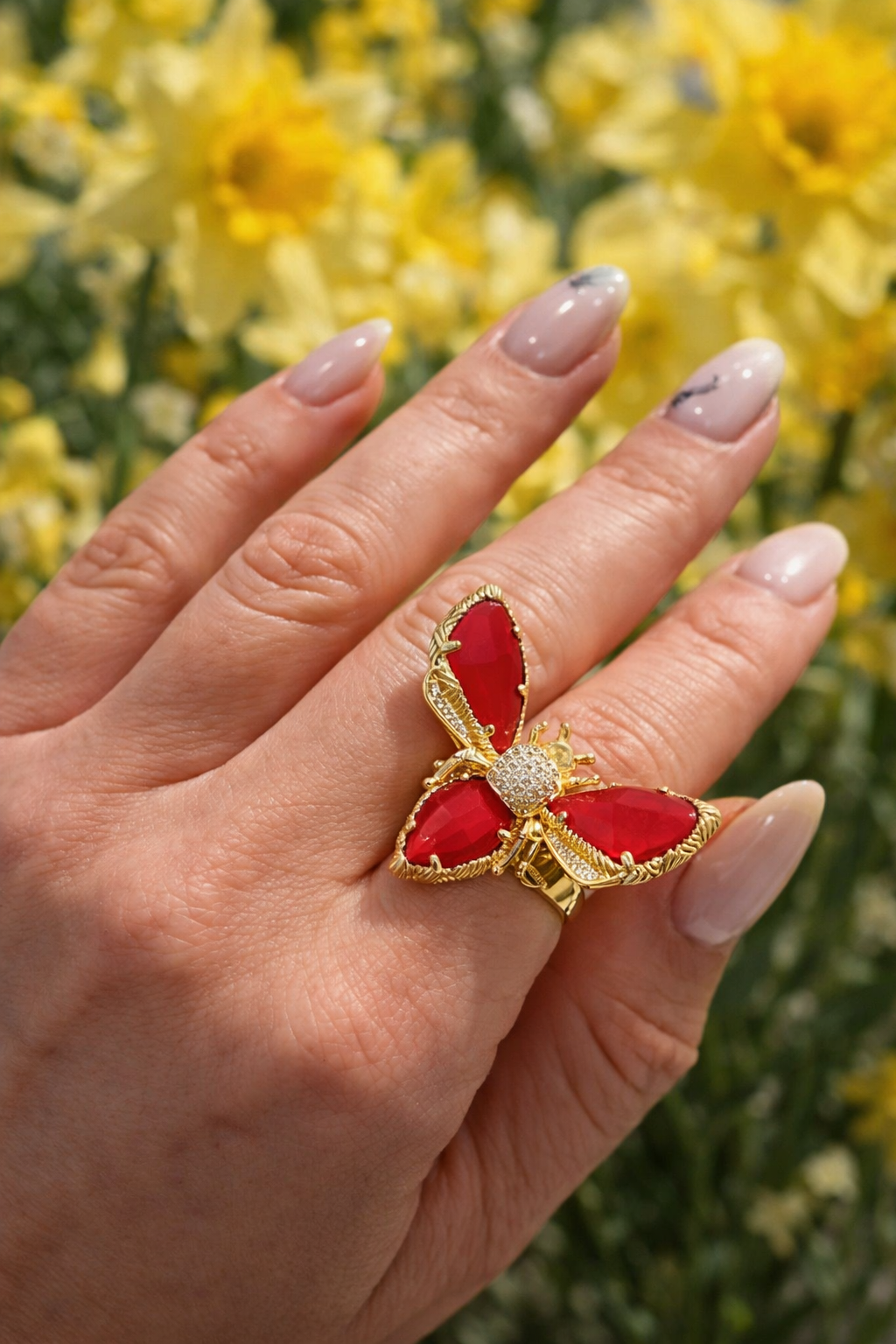 Tuvplāna fotogrāfija ar sievietes roku un dekoratīvu tauriņa gredzenu ar sarkaniem mirdzošiem akmeņiem zelta krāsas ietvarā uz izpludināta dzeltenu ziedu fona.