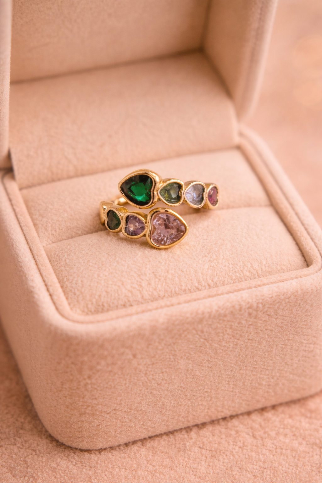 Zelta krāsas atvērts gredzens ar vairākiem dažādu krāsu kristāliem (zaļš, violets, rozā un gaiši zils), dažādās formās, novietots gaišā samta rotaslietu kastītē siltā apgaismojumā.