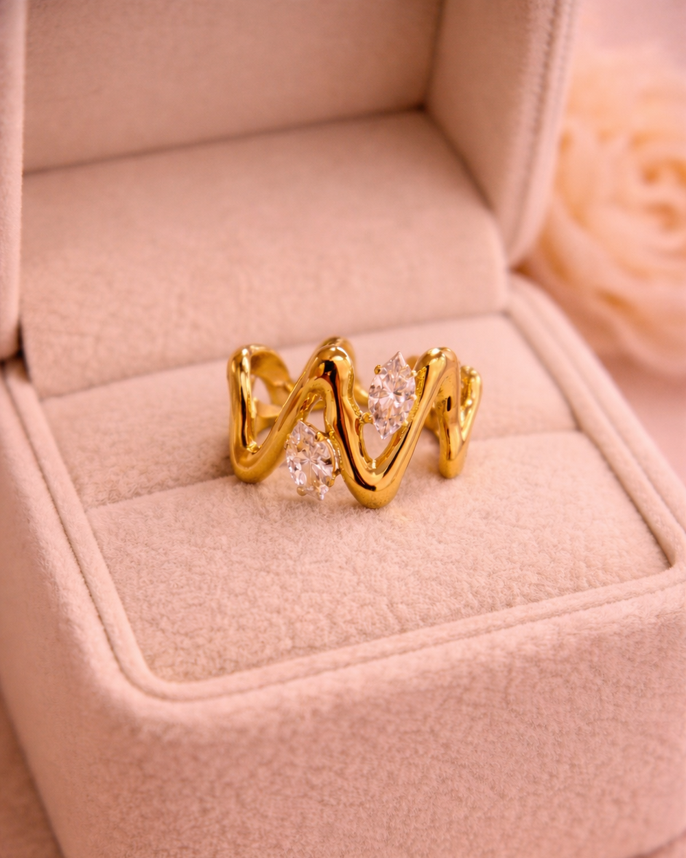 Zelta krāsas viļņveida dizaina gredzens ar diviem mirdzošiem caurspīdīgiem kristāliem, novietots uz bēšas samta rotaslietu kastītes siltā apgaismojumā.