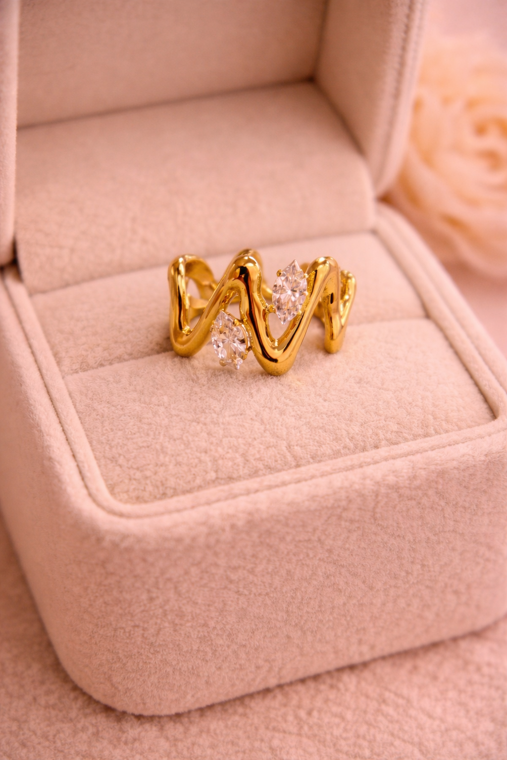 Zelta krāsas viļņveida dizaina gredzens ar diviem mirdzošiem caurspīdīgiem kristāliem, novietots uz bēšas samta rotaslietu kastītes siltā apgaismojumā.