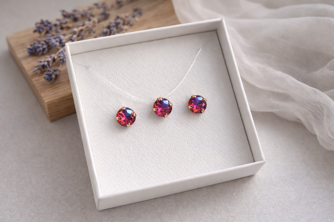 Minimālistiska rotu komplekta produktu fotogrāfija baltā kastītē – zeltītā ietvarā iestrādāti sarkani AB kristāli. Komplektā nagliņu auskari un kaklarota uz caurspīdīgas auklas. Fons gaišs ar koka dēlīti, žāvētu lavandu un viegli plīvojošu caurspīdīgu audumu, maigs dabīgais apgaismojums un sekls asuma dziļums.
