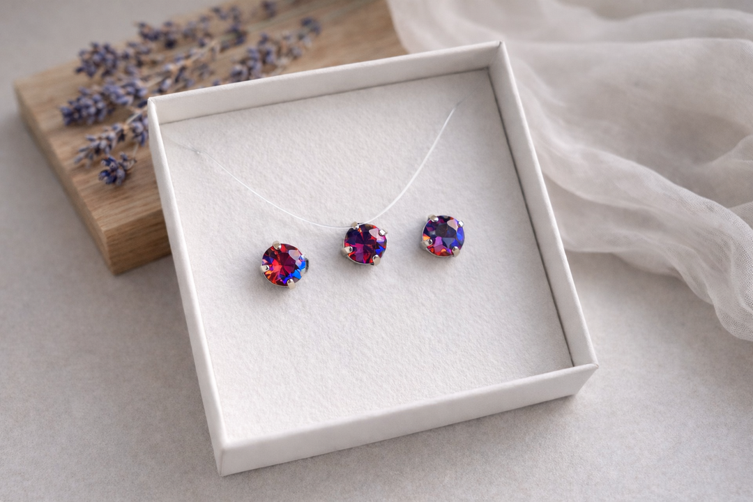 Minimālistiska rotu komplekta produktu fotogrāfija baltā kastītē – sudraba ietvarā iestrādāti sarkani kristāli ar izteiktiem ziliem atspīdumiem. Komplektā nagliņu auskari un kaklarota uz caurspīdīgas auklas. Fons gaišs, ar koka dēlīti, žāvētu lavandu un viegli izpludinātu audumu, maigs dabīgais apgaismojums.