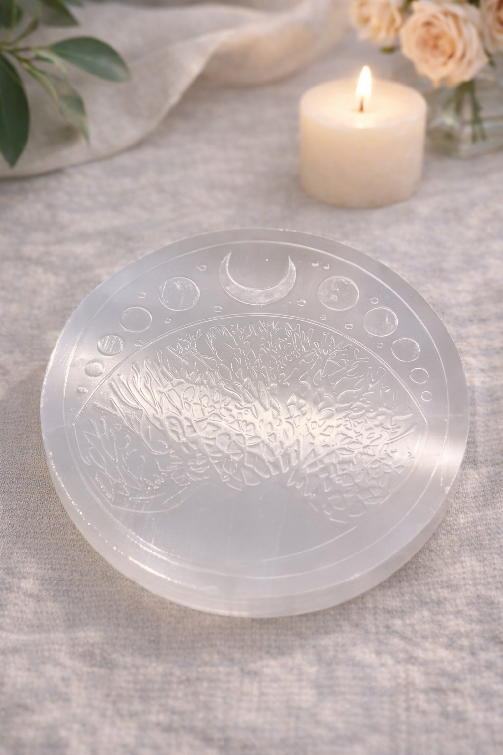 Selenīta attīrīšanas plātne ar iegravētu Dzīvības koku un Mēness fāzēm, novietota uz gaiša auduma fona dabīgā apgaismojumā.