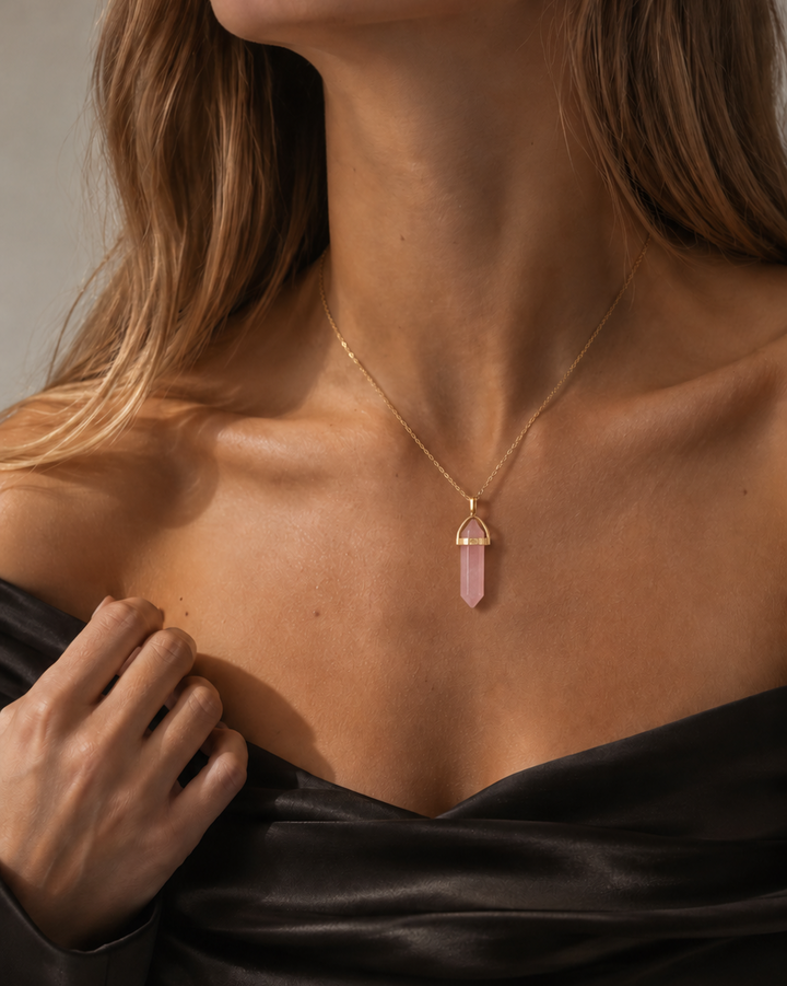 Elegants skaistuma portrets ar fokusu uz kaklu un atslēgas kauliem. Modele valkā melnu satīna off-shoulder topu un smalku dzeltenā zelta kaklarotu ar gaiši rozā rozā kvarca smailes formas kulonu.