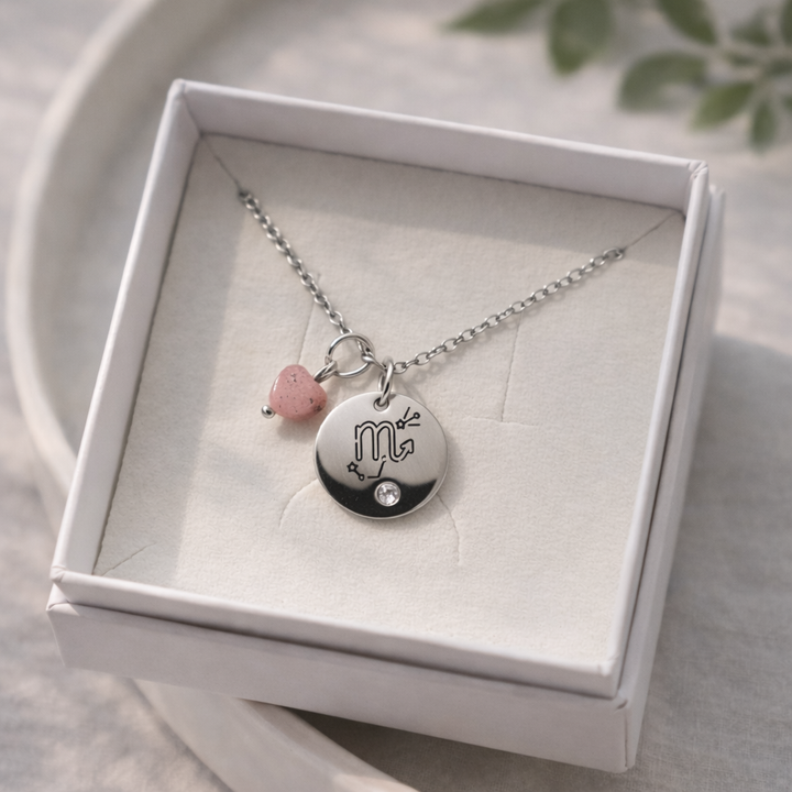 Sudraba kaklarota ar apaļu gravētu kulonu (Skorpiona simbols), nelielu kristālu un rozā akmens piekariņu, ievietota baltā rotaslietu kastītē uz gaiša keramikas šķīvja, ar maigu dabīgo apgaismojumu un izpludinātu zaļu augu fonā.