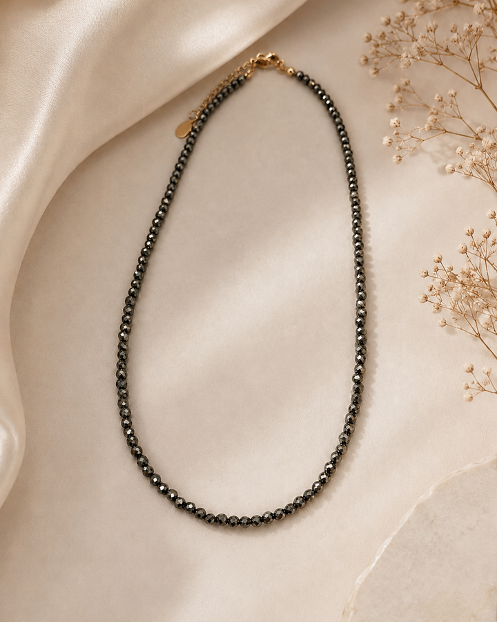 Smalka 3 mm pirīta kaklarota ar regulējamu zeltītu aizdari, eleganti novietota uz gaiša fona. Silta dabīgā gaisma izceļ pirīta akmeņu metālisko mirdzumu, bet zīdaina auduma un kaltētu ziedu akcenti rada minimālistisku, premium juvelierizstrādājumu estētiku.