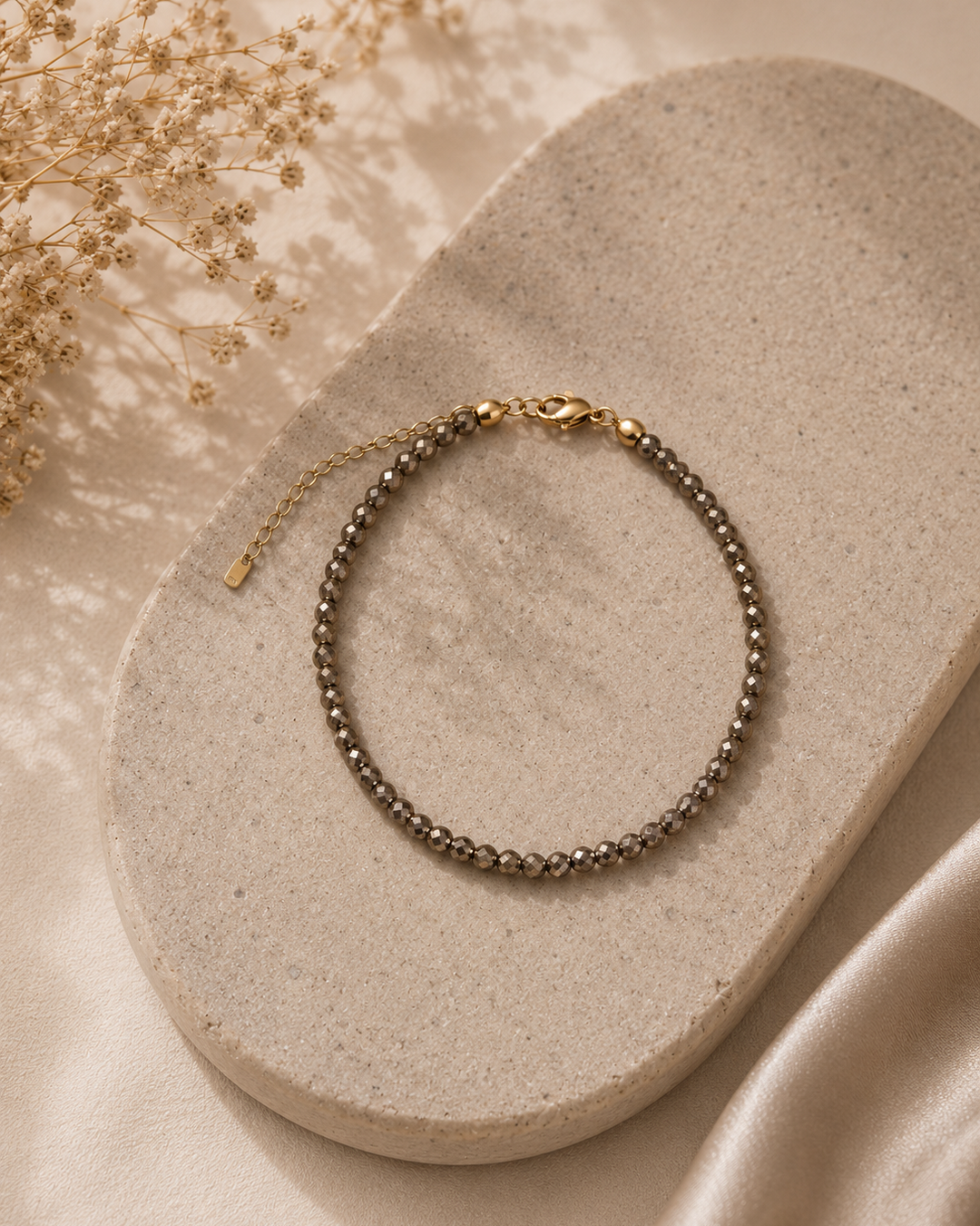 Smalka 3 mm pirīta rokassprādze ar zeltītu aizdari un pagarinājuma ķēdīti, eleganti novietota uz gaišas, ovālas akmens paplātes.