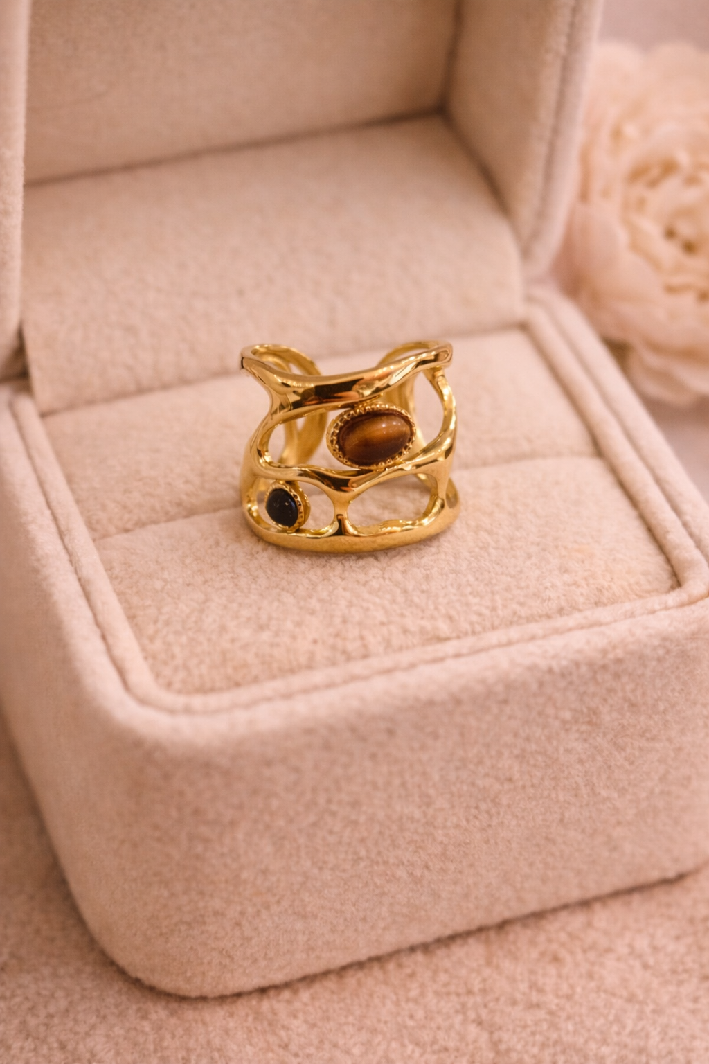 Zelta krāsas plata dizaina gredzens ar ažūra formu, dekorēts ar ovālu brūnu tīģeracs akmeni un mazu tumšu akcenta akmeni, novietots uz gaišas samta rotaslietu kastītes siltā apgaismojumā.