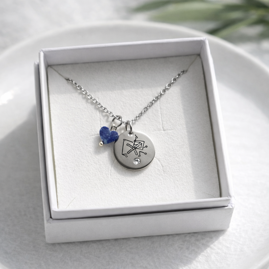 Sudraba kaklarota ar apaļu gravētu kulonu (Strēlnieka zodiaka simbols), nelielu kristālu un zilu akmens piekariņu, ievietota baltā rotaslietu kastītē uz gaiša keramikas šķīvja, ar maigu dabīgo apgaismojumu un izpludinātu zaļu augu fonā.