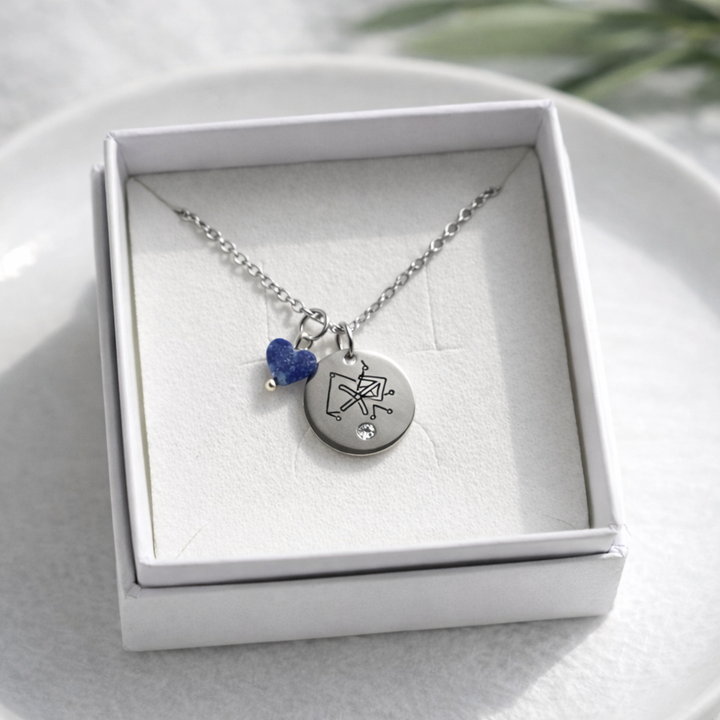 Sudraba kaklarota ar apaļu gravētu kulonu (Strēlnieka zodiaka simbols), nelielu kristālu un zilu akmens piekariņu, ievietota baltā rotaslietu kastītē uz gaiša keramikas šķīvja, ar maigu dabīgo apgaismojumu un izpludinātu zaļu augu fonā.