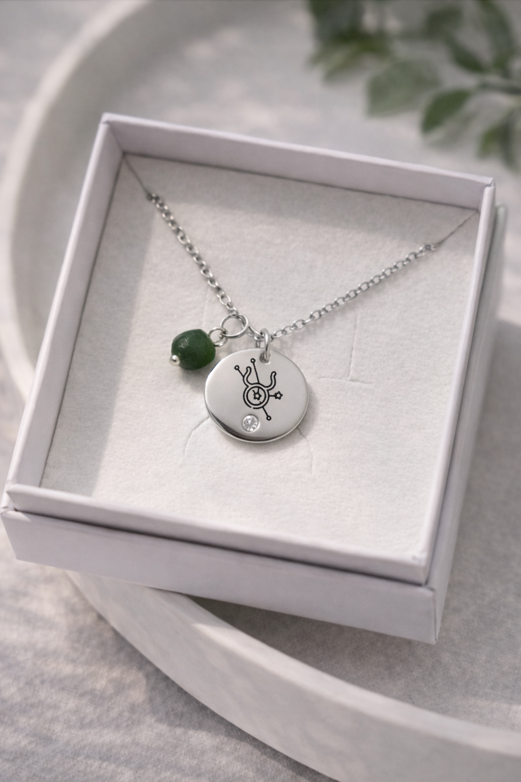 Sudraba kaklarota ar gravētu zodiaka simbola kulonu un zaļu akmens piekariņu, novietota baltā rotaslietu kastītē uz gaiša, dabīga fona ar maigu apgaismojumu.