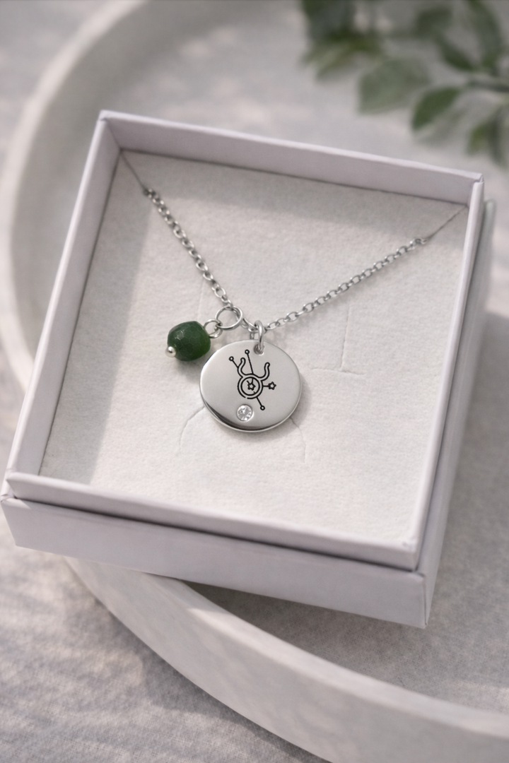 Sudraba kaklarota ar gravētu zodiaka simbola kulonu un zaļu akmens piekariņu, novietota baltā rotaslietu kastītē uz gaiša, dabīga fona ar maigu apgaismojumu.