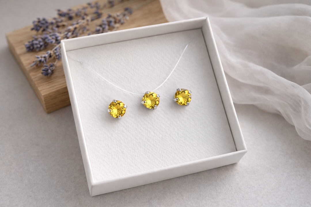 Minimālistiska rotu komplekta produktu fotogrāfija baltā kastītē – sudraba ietvarā iestrādāti dzelteni kristāli.Komplektā nagliņu auskari un kaklarota uz caurspīdīgas auklas. Fons gaišs, ar koka dēlīti, žāvētu lavandu un viegli izpludinātu audumu, maigs dabīgais apgaismojums.