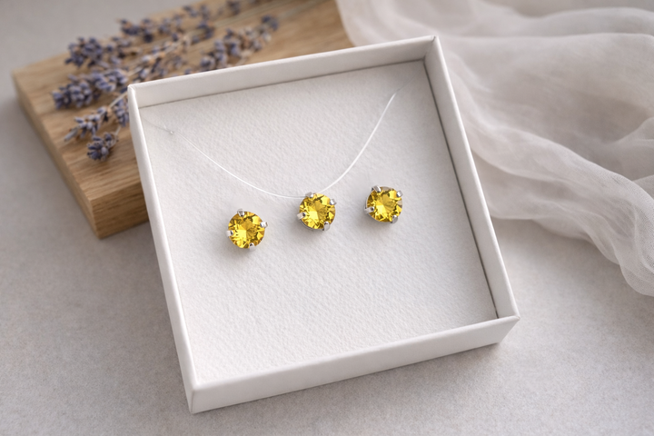 Minimālistiska rotu komplekta produktu fotogrāfija baltā kastītē – sudraba ietvarā iestrādāti dzelteni kristāli.Komplektā nagliņu auskari un kaklarota uz caurspīdīgas auklas. Fons gaišs, ar koka dēlīti, žāvētu lavandu un viegli izpludinātu audumu, maigs dabīgais apgaismojums.