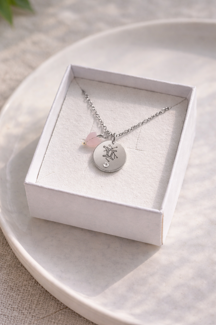 Sudraba kaklarota ar gravētu Dvīņu zodiaka kulonu un gaiši rozā akmens piekariņu, ievietota baltā rotaslietu kastītē uz gaiša, minimālistiska fona ar maigu dabīgo apgaismojumu.