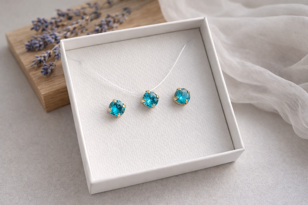 Minimālistiska rotu komplekta produktu fotogrāfija baltā kastītē – zeltītā ietvarā iestrādāti spilgti tirkīzzili kristāli. Komplektā nagliņu auskari un kaklarota uz caurspīdīgas auklas. Fons gaišs ar koka dēlīti, žāvētu lavandu un viegli plīvojošu caurspīdīgu audumu, maigs dabīgais apgaismojums un sekls asuma dziļums.
