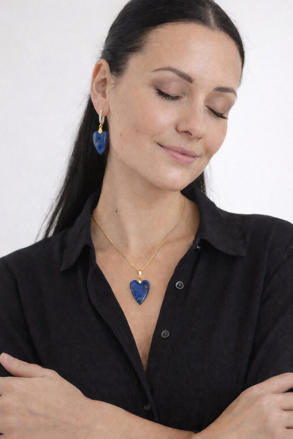 Sieviete ar aizvērtām acīm un vieglu smaidu, valkā zelta kaklarotu un auskarus ar lazurīta sirds formas kuloniem uz gaiša, minimālistiska fona.