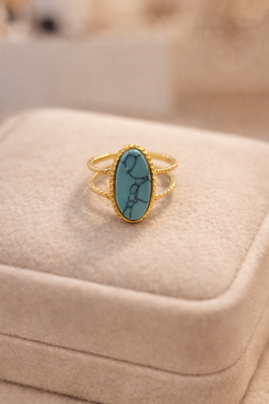 Zelta toņa gredzens ar ovālu zilu magnēzista akmeni un dubultu stīpiņas dizainu, novietots uz gaišas samta juvelierizstrādājumu kastītes maigā, siltā apgaismojumā.