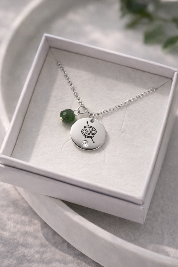 Sudraba kaklarota ar gravētu zodiaka kulonu un zaļu akmens piekariņu, ievietota baltā rotaslietu kastītē uz gaiša, minimālistiska fona ar dabīgu, maigu apgaismojumu.