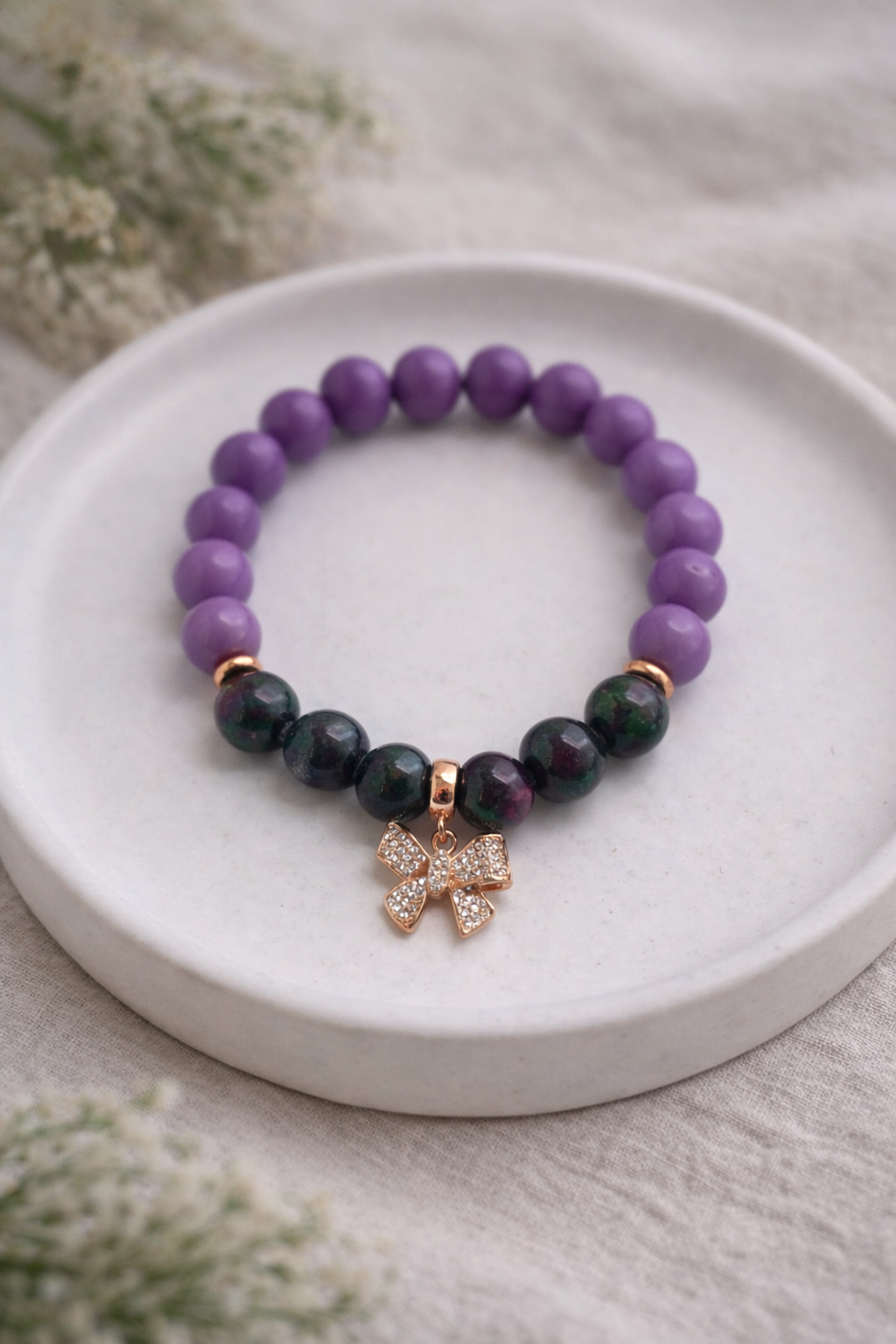 Minimālistiska produktu fotogrāfija ar žadeīta pērlīšu aproci, novietotu uz balta keramikas šķīvīša. Aproce veidota no gludām, apaļām zaļām žadeīta pērlēm ar zelta krāsas starpdetaļām un smalku piekariņu. Kompozīcija veidota uz neitrāla lina auduma, maigs dabīgais apgaismojums izceļ akmeņu faktūru un spīdumu.