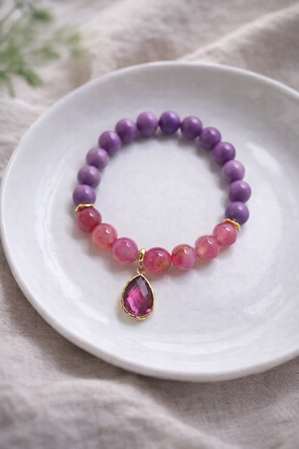 Dabīgo akmeņu aproce ar violetām un rozā pērlēm, simetriski izkārtota uz balta keramikas šķīvīša. Abās pusēs pa trim rozā akmens pērlēm, starp tām zelta krāsas starpdetaļas un centrā liels, slīpēts rozā kulons zelta ietvarā. Fons – gaišs lina audums ar maigi izpludinātu zaļu augu fonā, maiga dabīgā dienasgaisma.