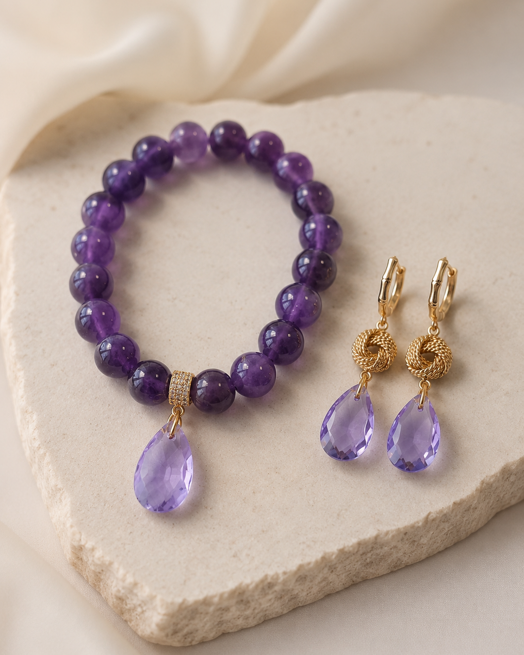 Elegants rotu komplekts uz gaišas akmens paplātes – rokassprādze no dabīgām ametista pērlēm ar violetu kristāla lāsīti un pieskaņoti zelta krāsas auskari ar violetiem kristāla piekariņiem.