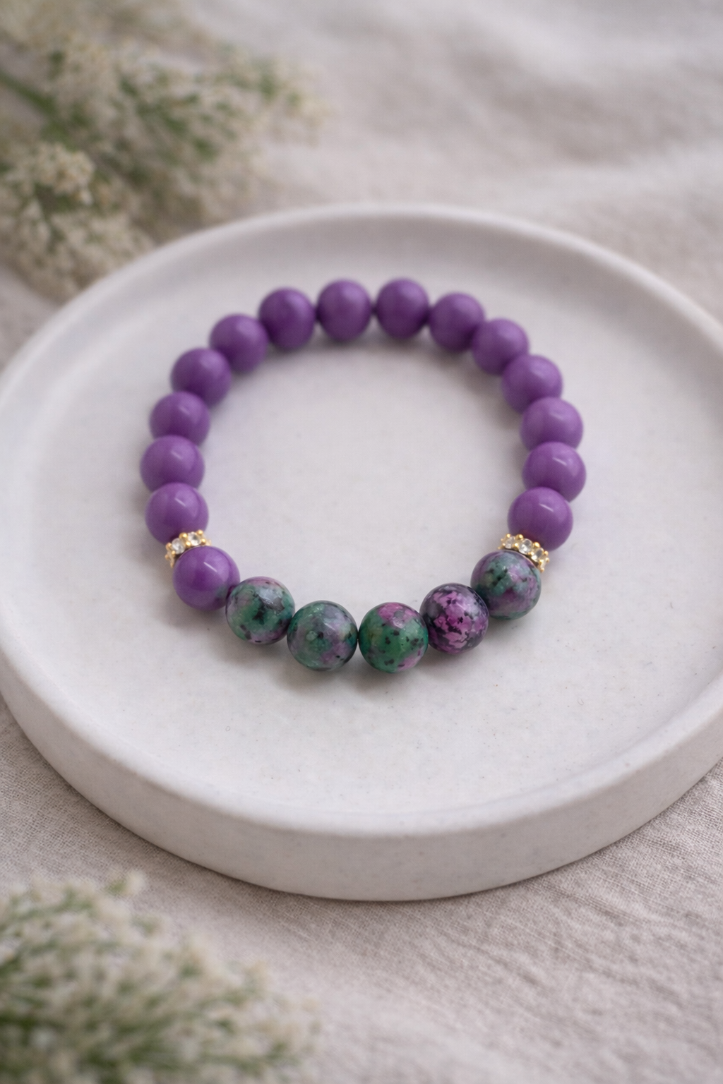 Minimālistiska produktu fotogrāfija ar dabīgo akmeņu aproci, novietotu uz balta keramikas šķīvīša. Aproce veidota no gludām violetām pērlēm un apaļiem zaļi-rozā rubīna zoisītā akmeņiem, papildināta ar zelta krāsas starpdetaļām ar smalkiem kristāliem. Kompozīcija izvietota uz gaiša lina auduma ar maigi izpludinātiem zaļiem augu elementiem fonā, mīkstā dabīgā apgaismojumā.