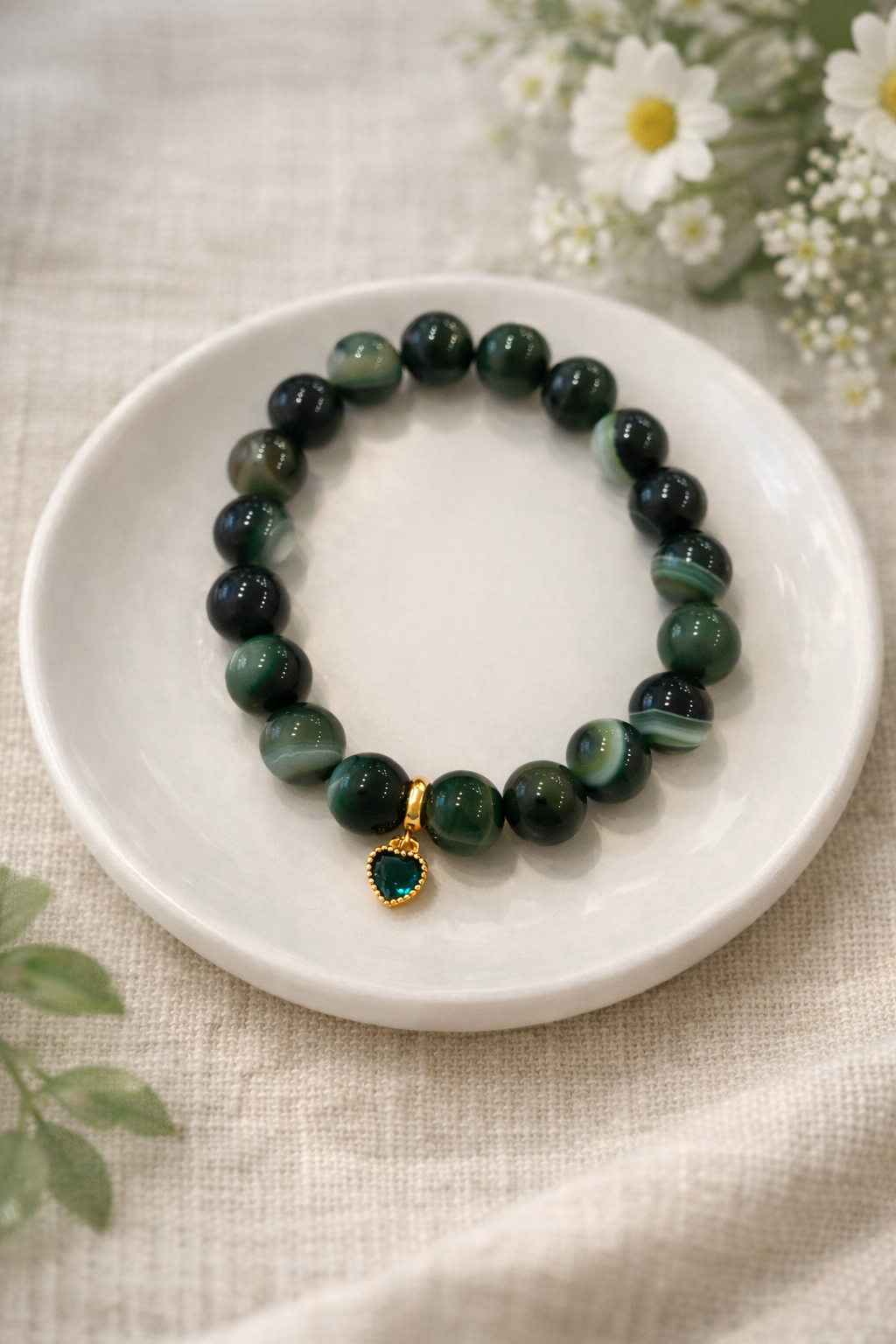 Zaļa akmens pērlīšu rokassprādze ar zelta sirds piekariņu, novietota uz balta keramikas šķīvīša uz gaiša lina auduma, fonā maigi izpludināti balti ziedi un zaļi augi.