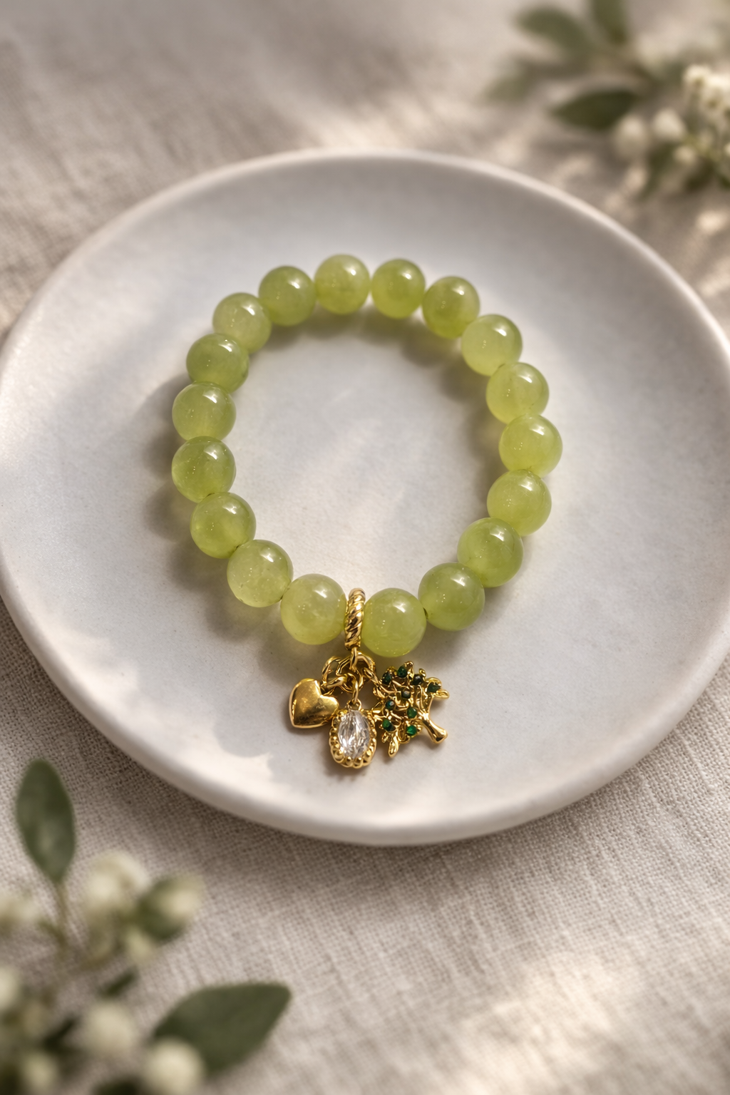 Minimālistiska gaiši zaļu dabīgo akmeņu aproce ar spīdīgām pērlēm, novietota uz balta keramikas šķīvīša. Aprocei piekariņi zelta tonī – sirds, caurspīdīgs kristāls un koka motīvs ar zaļiem akcentiem. Fons – gaišs lina audums ar maigi izpludinātiem zaļiem augu elementiem un maigu dabīgo apgaismojumu.