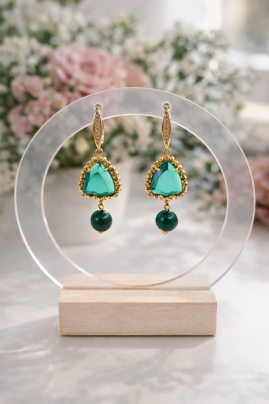 elta krāsas auskari ar zaļu trijstūra formas akmeni un tumši zaļu lodītes piekariņu, eleganti novietoti uz caurspīdīga rotu statīva ar koka pamatni. Fonā maigi izpludināti gaiši ziedi, radot romantisku un mājīgu produktu fotogrāfijas noskaņu.