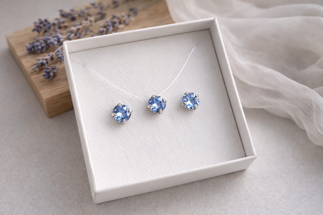 Minimālistiska rotu komplekta produktu fotogrāfija baltā kastītē – sudraba ietvarā iestrādāti sarkani kristāli ar izteiktiem ziliem atspīdumiem. Komplektā nagliņu auskari un kaklarota uz caurspīdīgas auklas. Fons gaišs, ar koka dēlīti, žāvētu lavandu un viegli izpludinātu audumu, maigs dabīgais apgaismojums.