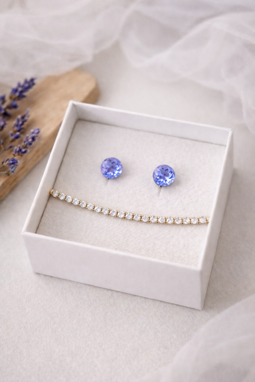 Balta rotaslietu kastīte ar baltu samta iekšpusi, kurā izvietoti apaļi violeti mirdzoši auskari un smalka zelta tenisa stila rokassprādze ar dzidriem akmentiņiem; fonā gaiša matēta virsma, koka dēlītis ar žāvētu lavandu un viegli izpludināts caurspīdīgs audums.