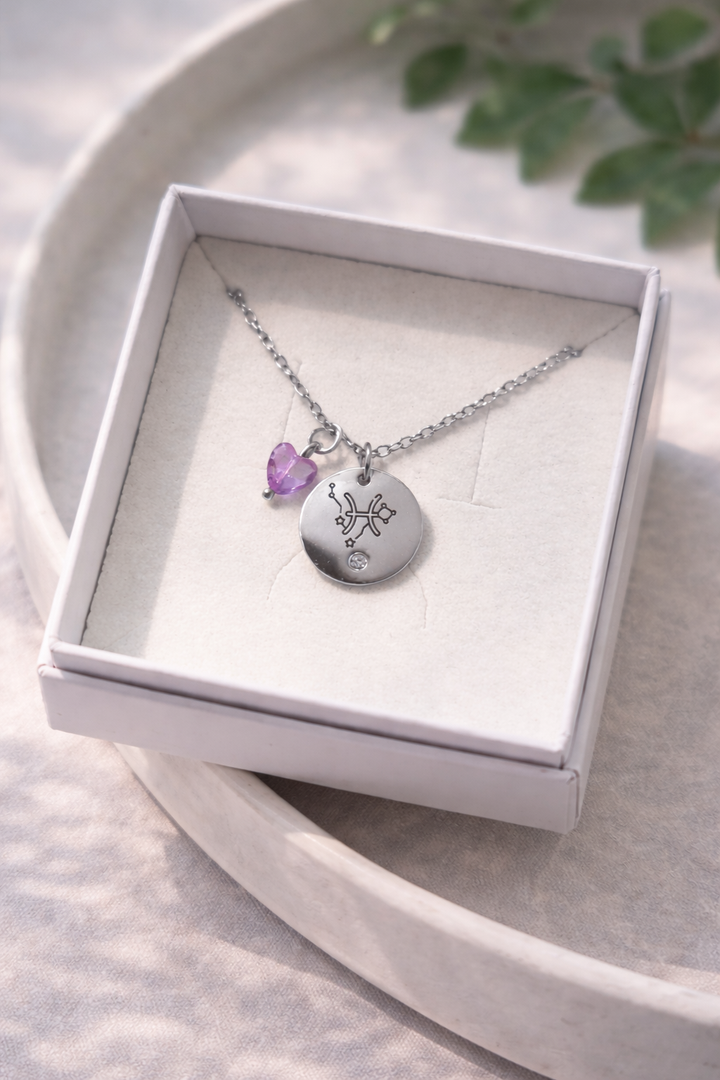 Sudraba kaklarota ar gravētu zodiaka kulonu un violetu sirds formas akmens piekariņu, ievietota baltā rotaslietu kastītē uz gaiša, dabīga fona ar maigu apgaismojumu.